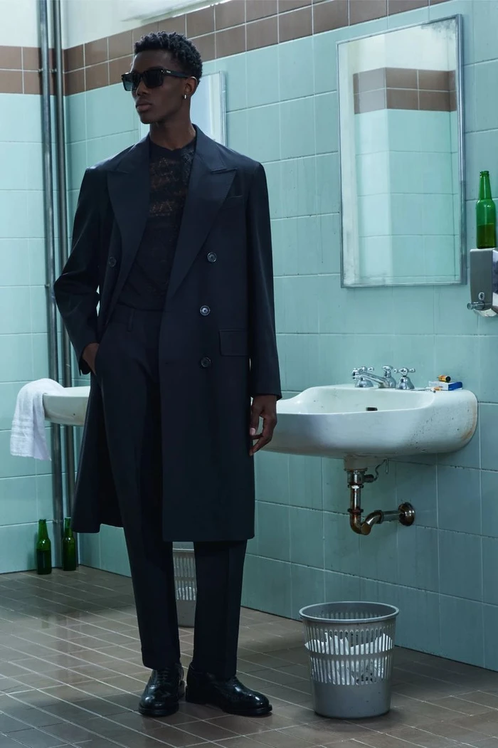 Model in dark coat and sunglasses standing in a tiled restroom with sinks and a mirror for Dsquared2 Resort 2026 lookbook.