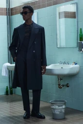 Model in dark coat and sunglasses standing in a tiled restroom with sinks and a mirror for Dsquared2 Resort 2026 lookbook.