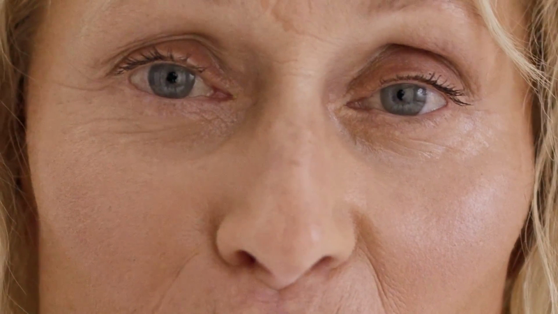 Close-up portrait of a mature woman's face featuring her blue eyes, fair skin, and blonde hair, showcasing natural skin textures and details.