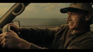 Man wearing a baseball cap driving a car with coastal scenery visible through the window
