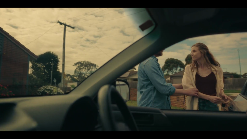 A man and woman stand outside of a car, viewed through the windshield. The suburban background includes houses and a cloudy sky.
