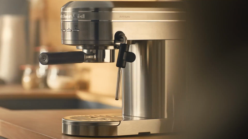 Stainless steel KitchenAid Artisan espresso machine on a wood countertop in a kitchen setting. Product shot focuses on machine details.
