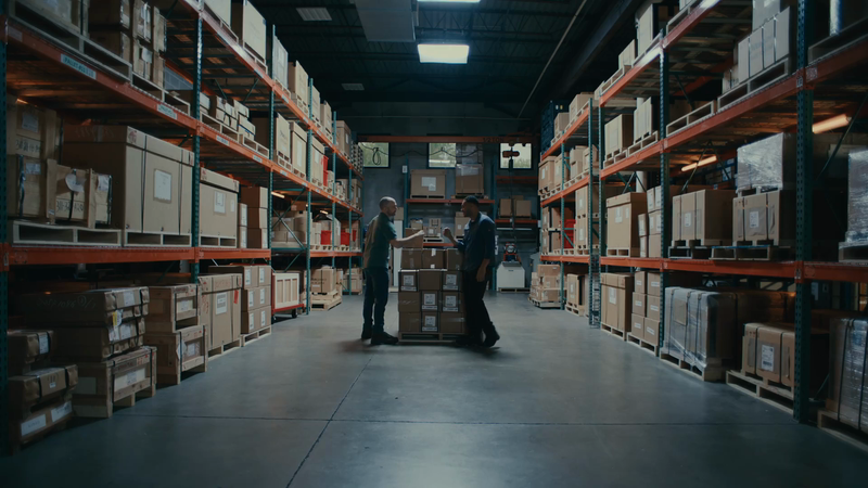 Two adult men face each other in a warehouse, stacked boxes on shelves around them in an indoor commercial storage setting.