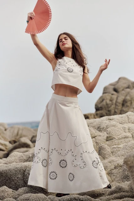 A woman in a white two-piece outfit stands on rocks near the ocean holding a fan, showcasing fashion, lifestyle, and outdoor elements.