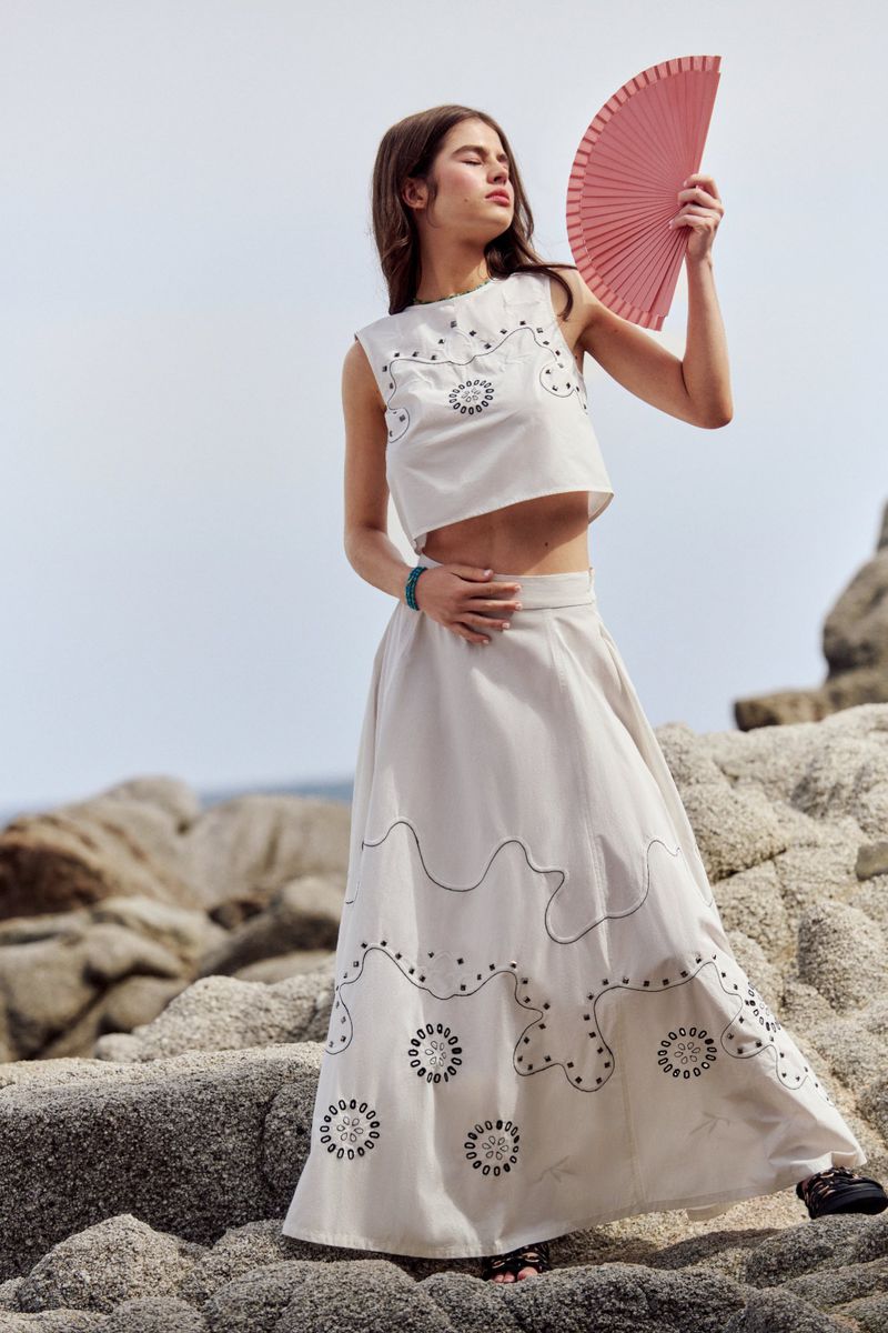 Woman in a white embroidered crop top and long skirt holding a pink fan, posing on coastal rocks against a bright sky backdrop.