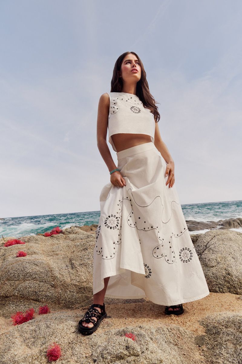 Woman in a two-piece dress with circular patterns stands on a rock near the ocean with the sky in the background.