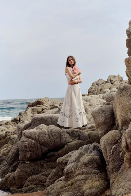Young woman in a white dress poses on a rocky coast with ocean in the background on a cloudy day, fashion photography.