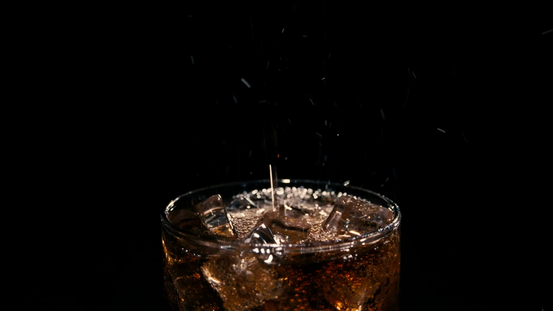 Close-up shot of a glass filled with dark cola and ice cubes with liquid splashing against a dark background.