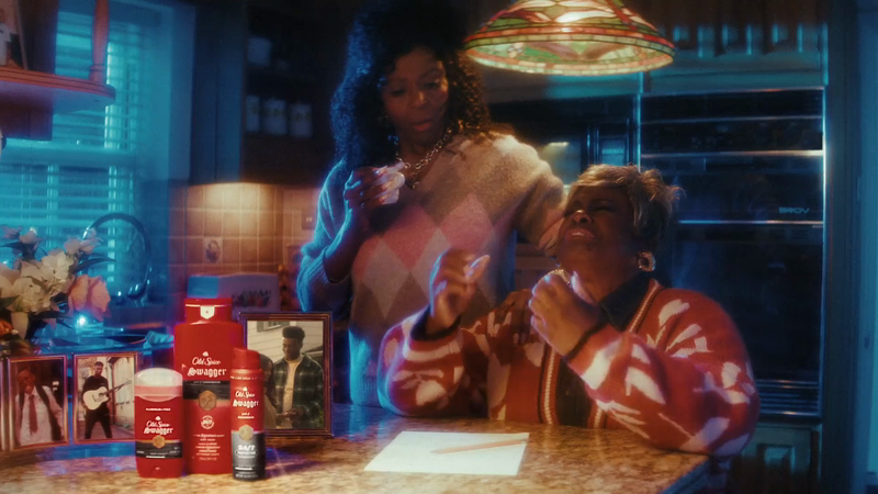 Two women seated at a kitchen counter with Old Spice Swagger grooming products, framed photos, and a cozy, vintage-style setting.