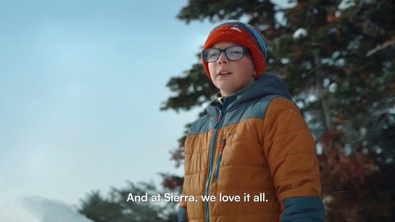 Boy in snow wearing hat and glasses with tan and blue coat in wintery outdoor setting, evergreen trees in background, close-up shot.