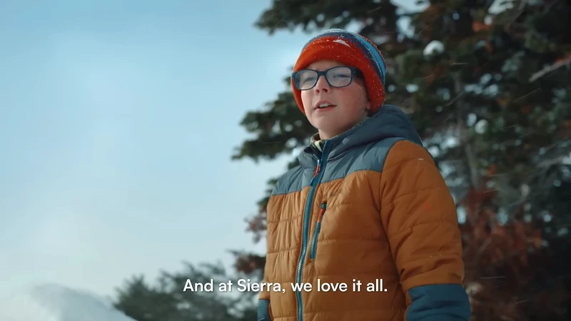Boy in snow wearing hat and glasses with tan and blue coat in wintery outdoor setting, evergreen trees in background, close-up shot.