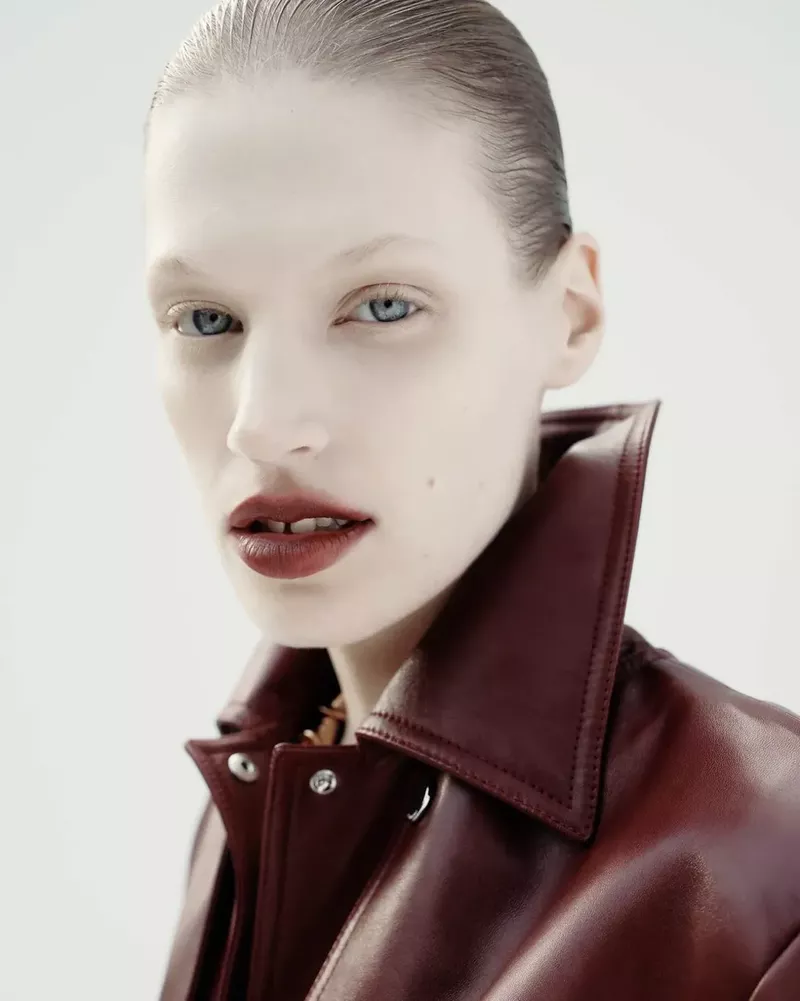 Close-up portrait of a model with slicked-back hair wearing a red leather jacket against a white background in a studio setting.