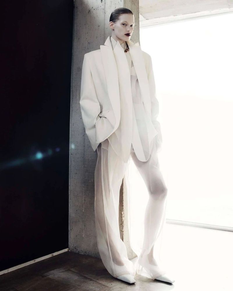 Fashion shot of a fair-skinned adult female model in a white jacket and sheer pants standing indoors against a concrete column.