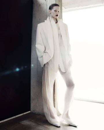 Fashion shot of a fair-skinned adult female model in a white jacket and sheer pants standing indoors against a concrete column.