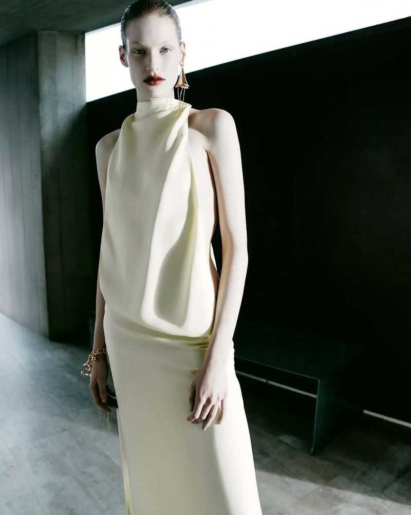 Elegant female fashion model in long flowing cream dress, gold jewelry, and striking red lipstick against dark architectural background.