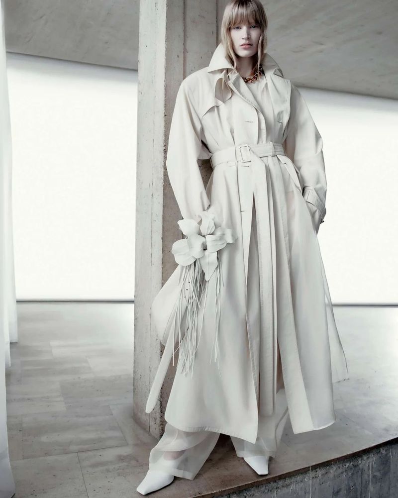 A woman in a white trench coat and pants poses indoors, holding a floral bouquet, in a fashion shot with concrete columns and natural light.