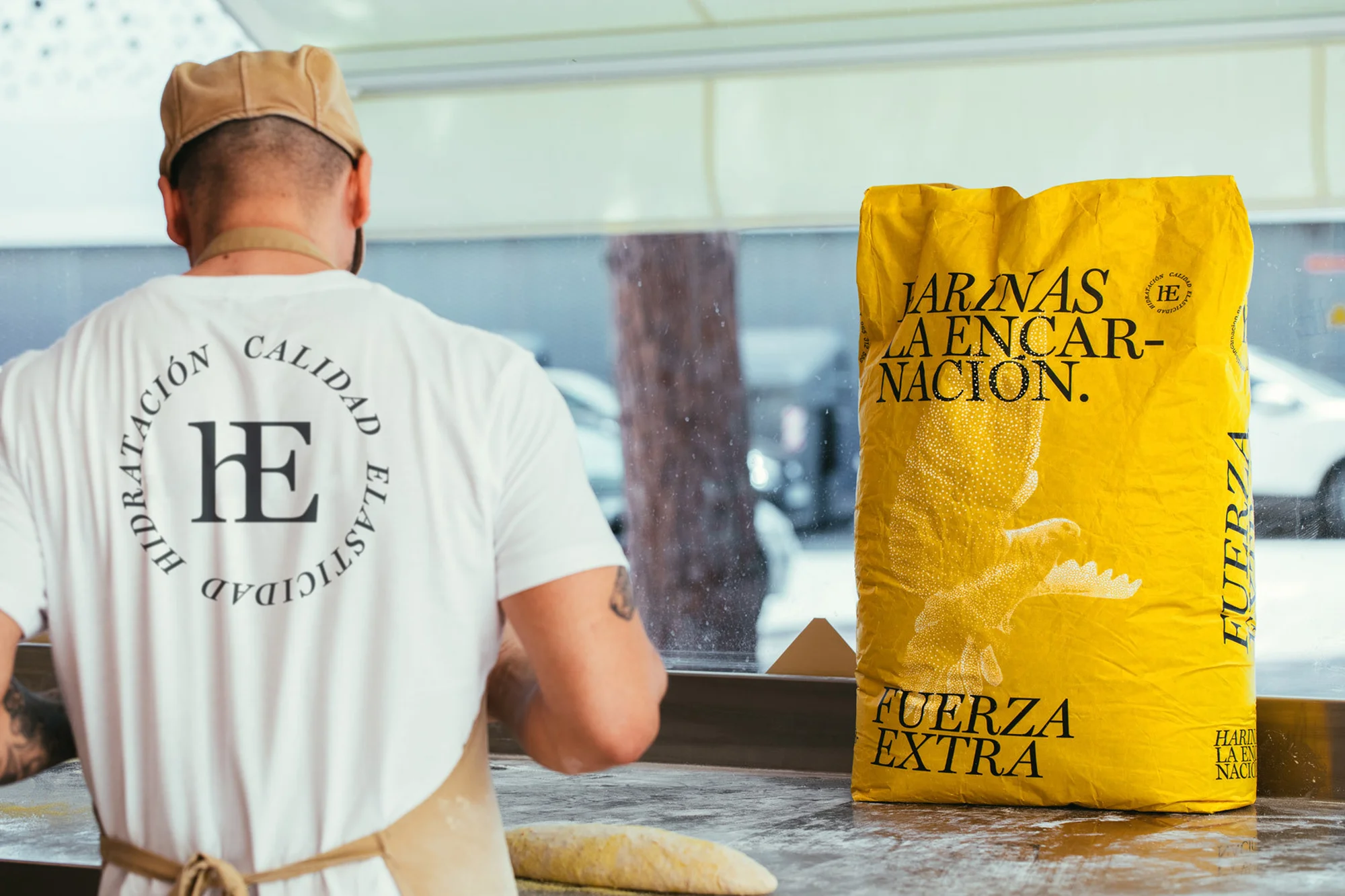 A baker wearing a white t-shirt is shown from the back next to a yellow bag of flour, preparing dough at a bakery counter.
