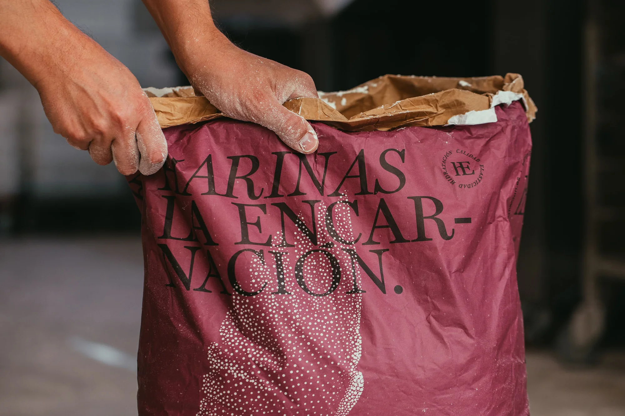 Hands opening a maroon paper bag of 'Harinas La Encarnacion' flour, with white dotted pattern near bottom, neutral background.