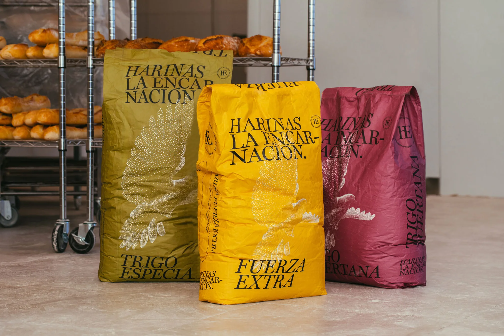 Three colorful bags of flour stand side by side in a bakery, advertising 'Harinas La Encarnacion' with an illustration of wheat.