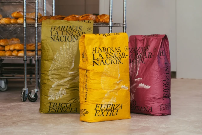 Three colorful bags of flour stand side by side in a bakery, advertising 'Harinas La Encarnacion' with an illustration of wheat.