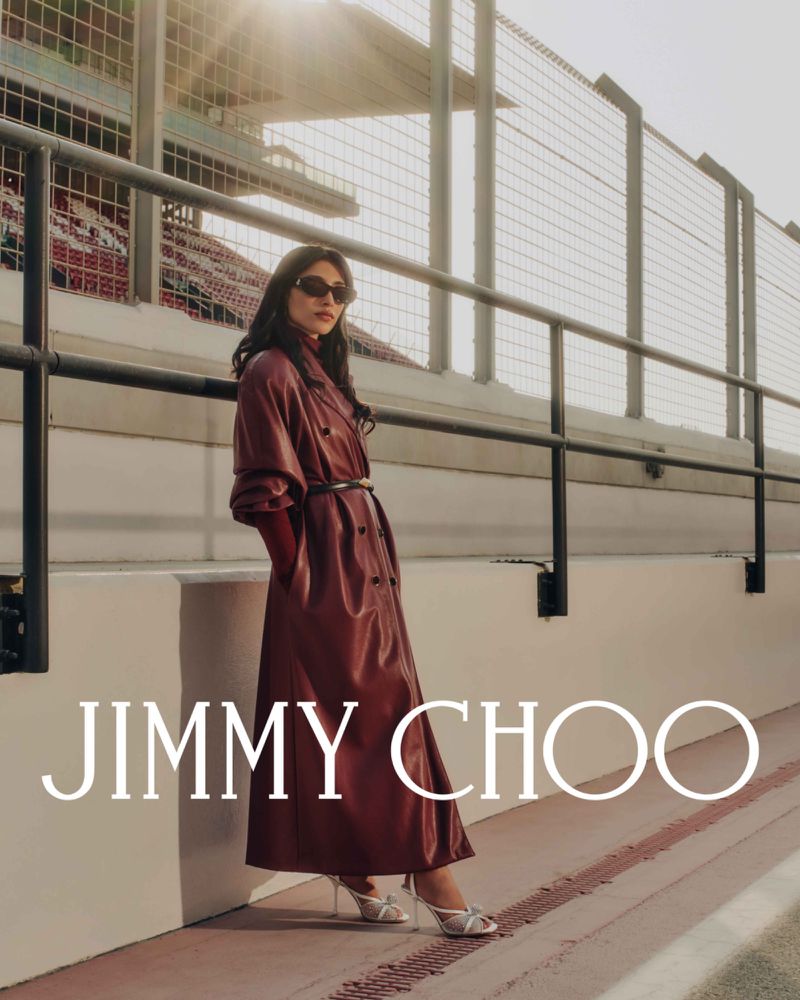 A woman in a maroon trench coat poses outdoors next to a white retaining wall, with metal fencing and light flares in the background.