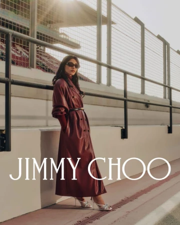 A woman in a maroon trench coat poses outdoors next to a white retaining wall, with metal fencing and light flares in the background.