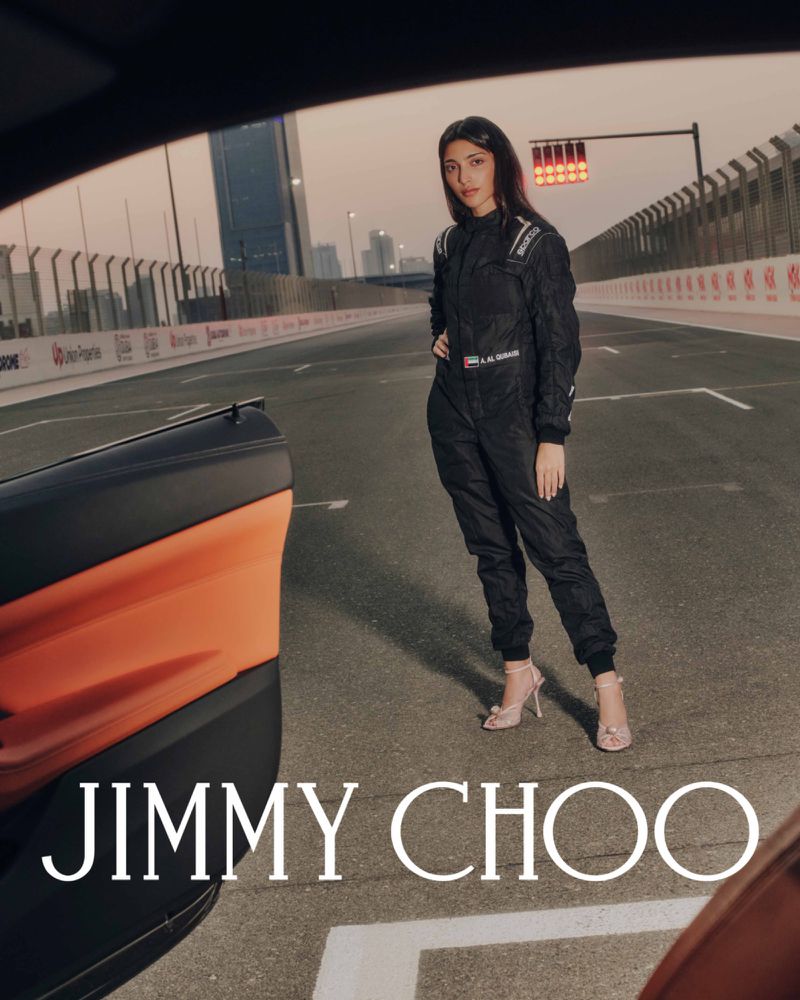 Woman in black racing jumpsuit poses on race track with orange sports car and Jimmy Choo logo in the foreground at dusk.