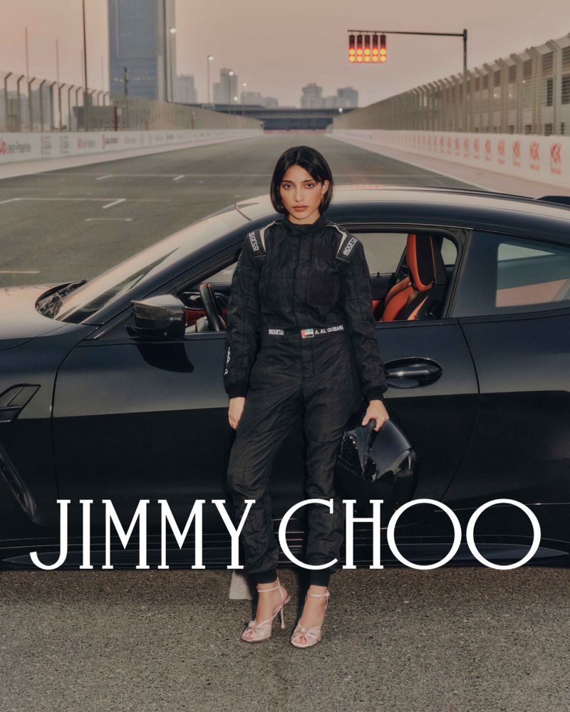 A woman in a racing suit stands in front of a black sports car on a racetrack at dusk with city buildings in the background.