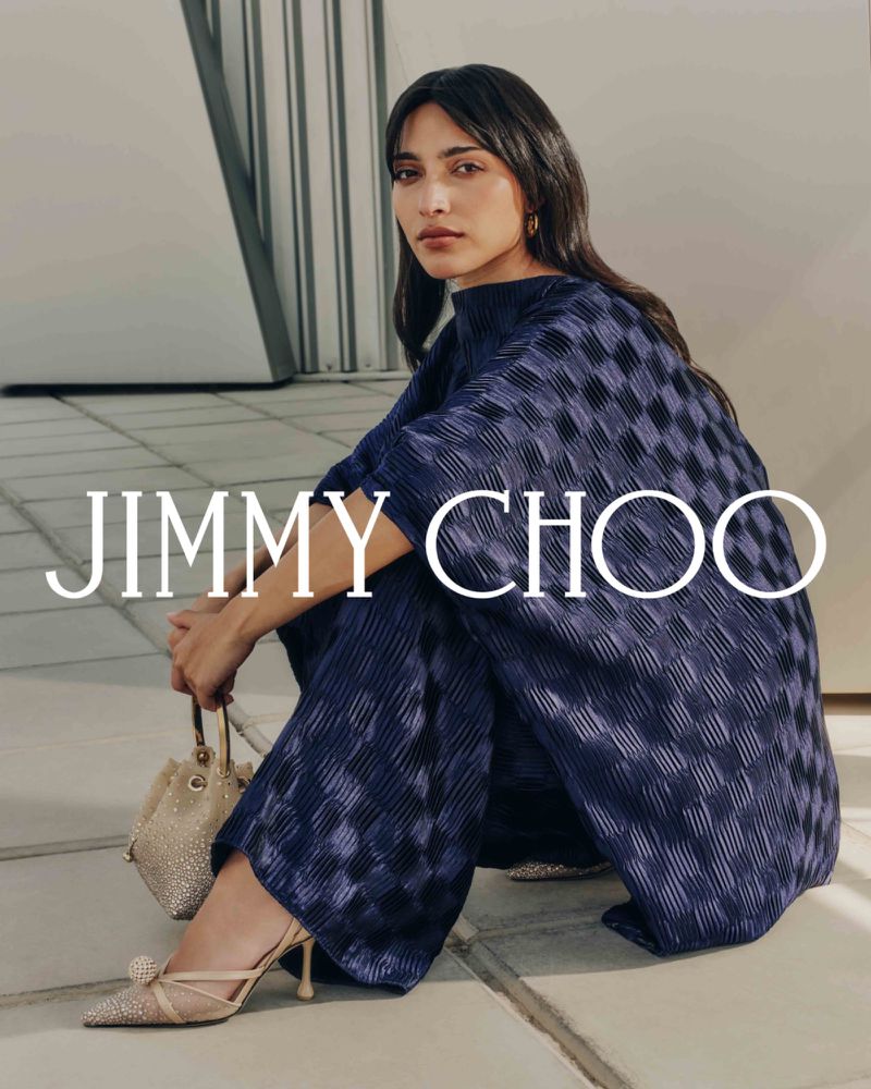 Brunette woman in a dark blue checkered two-piece outfit sits in a studio holding a small bag and wearing heels from Jimmy Choo.
