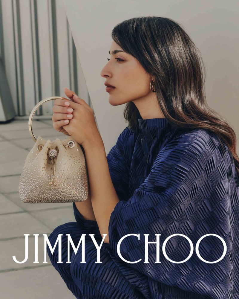 Elegant woman in blue dress sits near a white surface, holding a light-gold bucket bag with metallic details, and the Jimmy Choo logo.