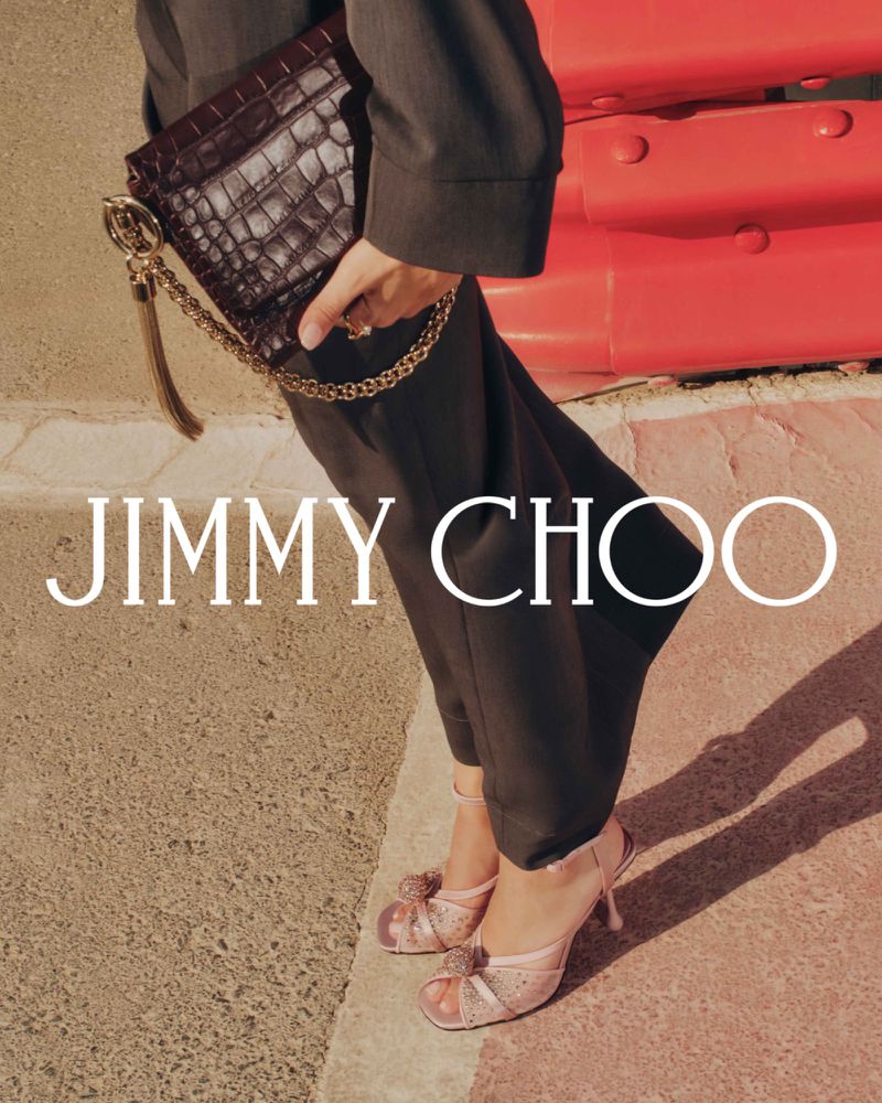 Fashion shot featuring a woman holding a dark brown crocodile-patterned clutch with gold chain, paired with pink high-heeled shoes outdoors.