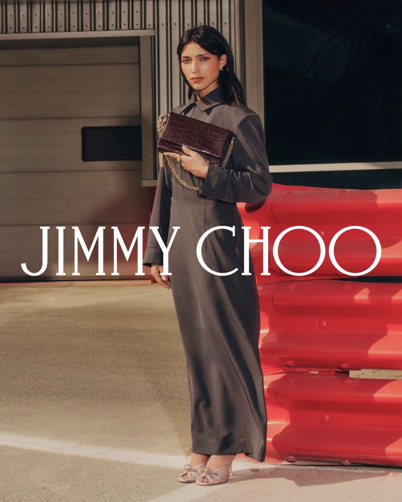 Fashion shot of an adult woman wearing a full-length gray dress and holding a burgundy crocodile print purse near a red barrier.