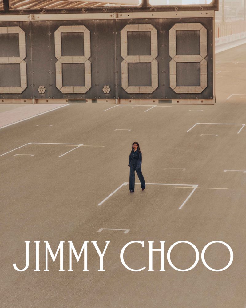 Woman in jumpsuit stands on court with large digital scoreboard and Jimmy Choo logo. Fashion shot with sepia tone.