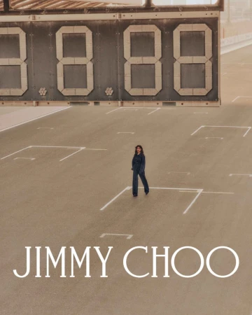 Woman in jumpsuit stands on court with large digital scoreboard and Jimmy Choo logo. Fashion shot with sepia tone.