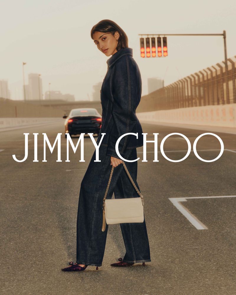 A woman in a denim jumpsuit and heels carries a handbag while standing on a street with a car and traffic lights in the background.
