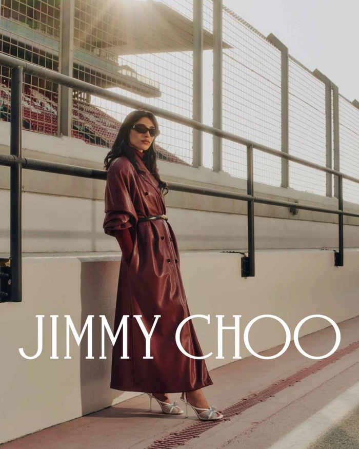 Woman in burgundy leather trench coat and silver heels leaning against a wall outdoors, featuring the Jimmy Choo brand overlay