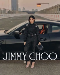 Woman in a black racing jumpsuit and heeled sandals standing beside a parked black sports car on a racetrack.