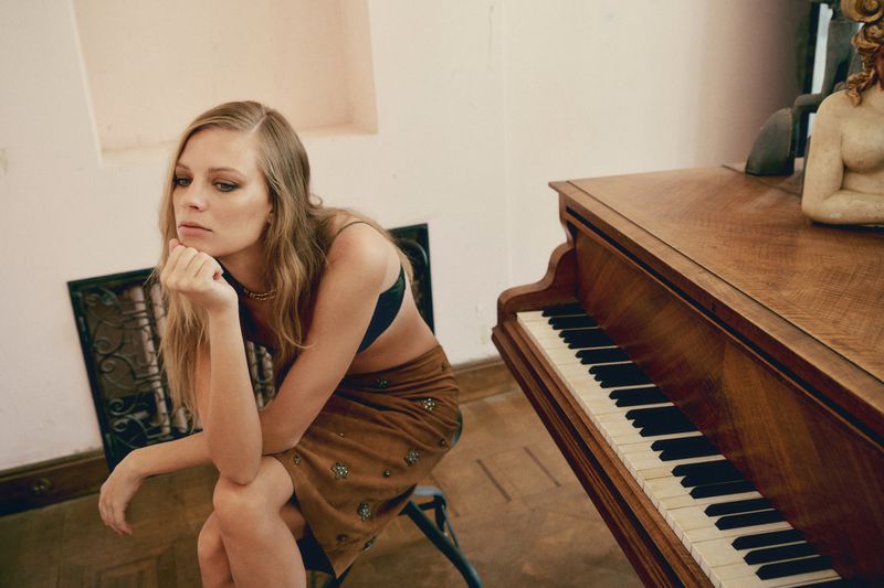 Indoor portrait of a blond woman sitting next to a piano in a skirt and bralette, with a statue in the background.