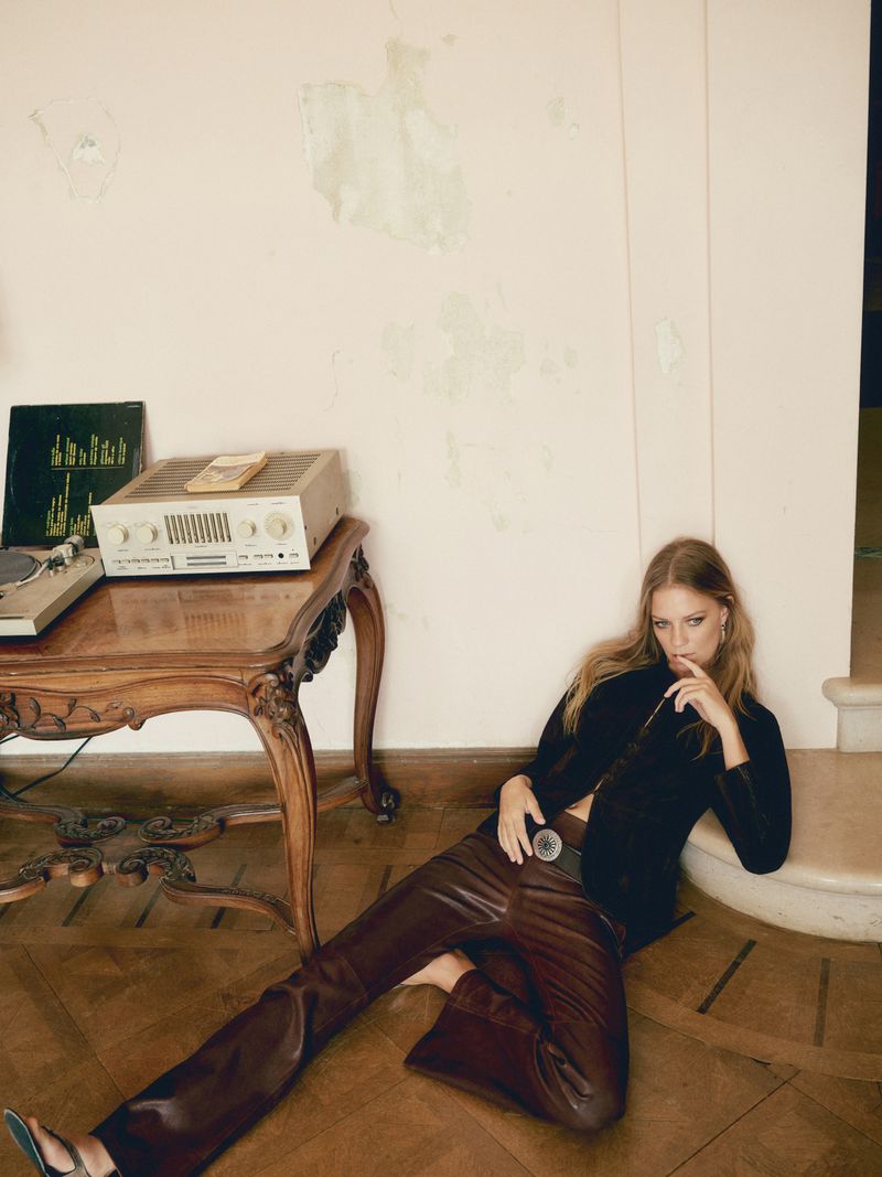 A woman in brown pants and a black jacket sits on a wooden floor in front of a wooden table with vintage audio equipment and vinyl records.