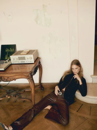 A woman in brown pants and a black jacket sits on a wooden floor in front of a wooden table with vintage audio equipment and vinyl records.