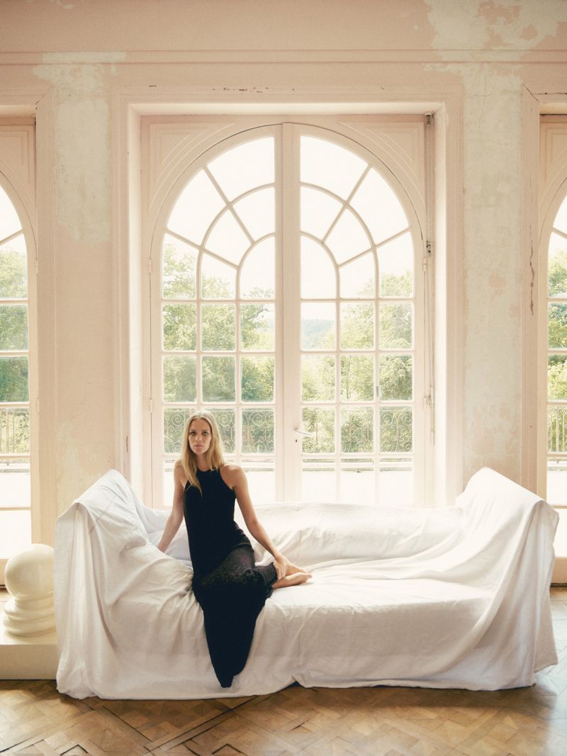 A young woman in a black dress sits on a white-draped couch in a room with large arched windows letting in daylight, posing for a fashion portrait.
