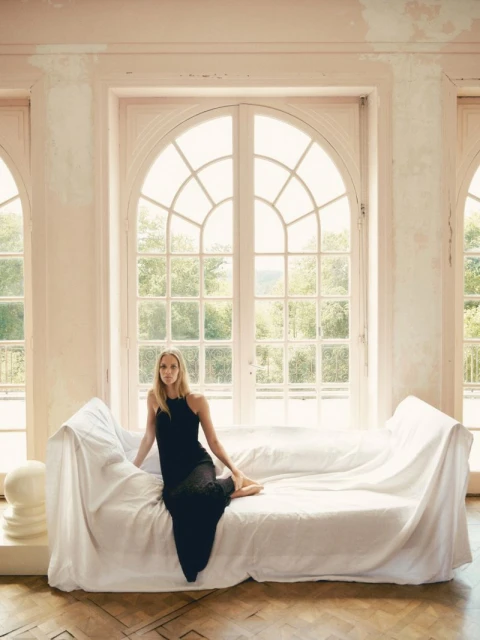 A young woman in a black dress sits on a white-draped couch in a room with large arched windows letting in daylight, posing for a fashion portrait.