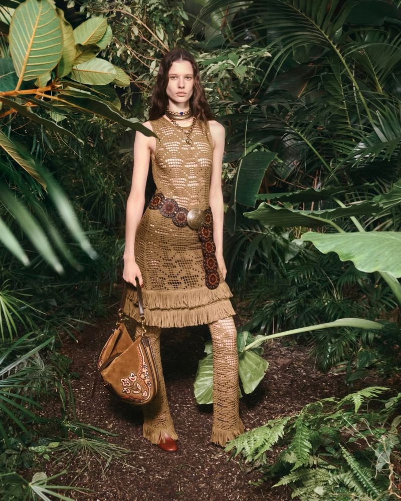 A female fashion model poses in a leafy, wooded setting, wearing a tan crocheted dress, crocheted leggings, a belt, necklace, and carrying a suede bag.