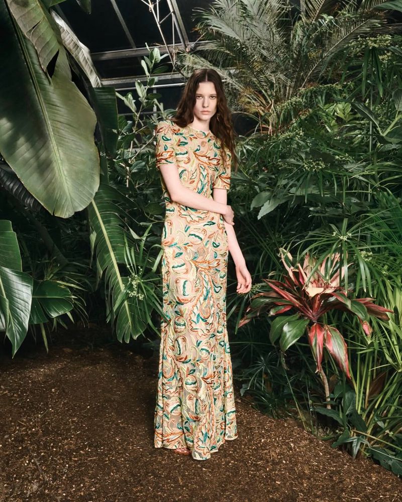 A young adult woman poses in a full-length gown with a floral pattern in a lush indoor botanical garden filled with greenery.
