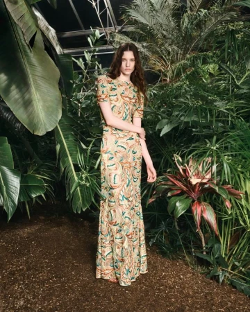 A young adult woman poses in a full-length gown with a floral pattern in a lush indoor botanical garden filled with greenery.