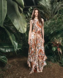 Model standing on a dirt path surrounded by lush tropical palm leaves and foliage wearing a patterned paisley maxi dress.