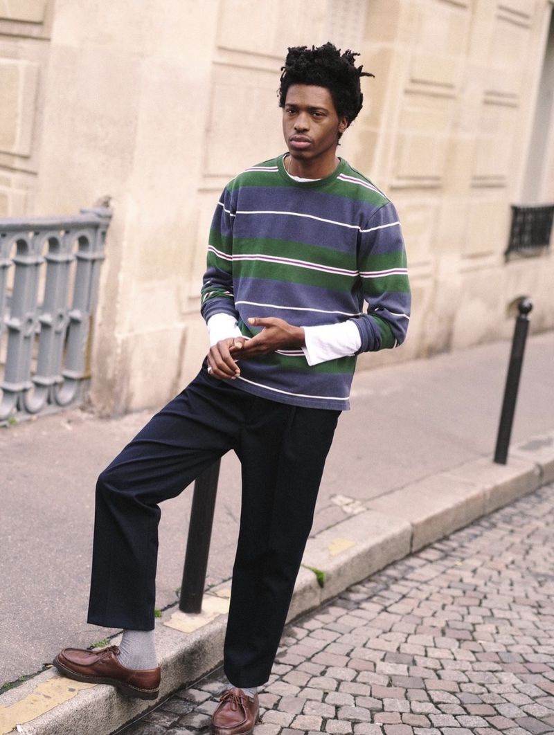 Adult male in striped shirt and navy trousers standing on city street with cobblestone, leaning against wall and metal railing outdoors.