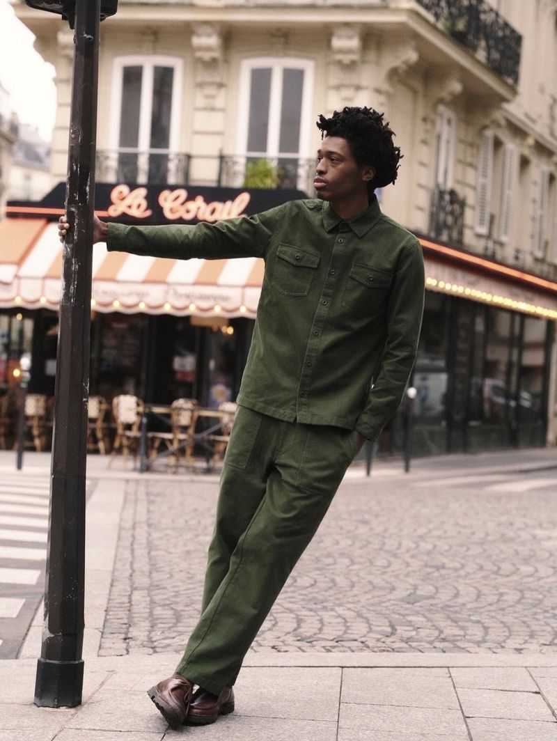 Young man in olive green suit leans on street lamp in Paris, France, with cobblestone street and building in the background.