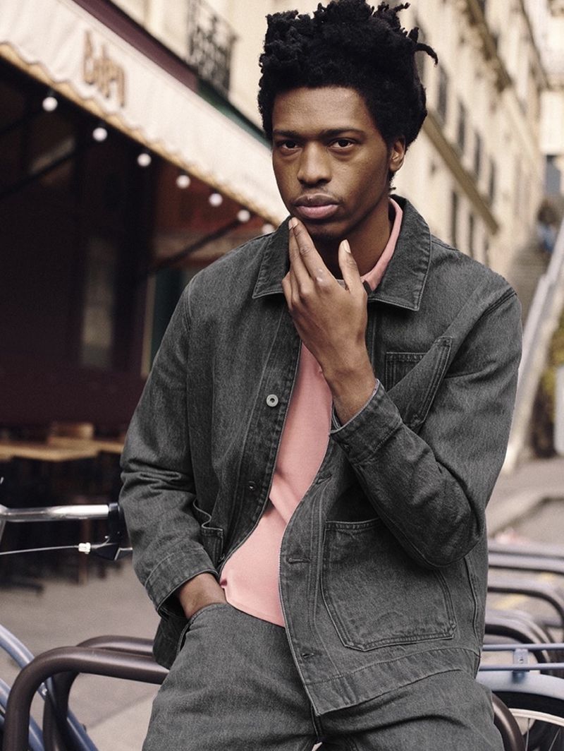 An adult male in a denim jacket and pants poses in an urban setting with a building and railings visible in the background.