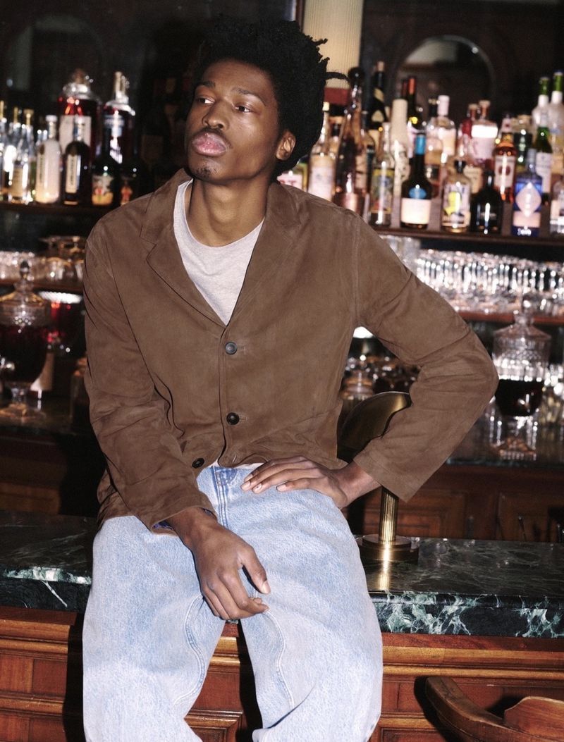 A young Black man sits on a bar counter in a brown jacket and light blue jeans, exuding casual style in an indoor setting.